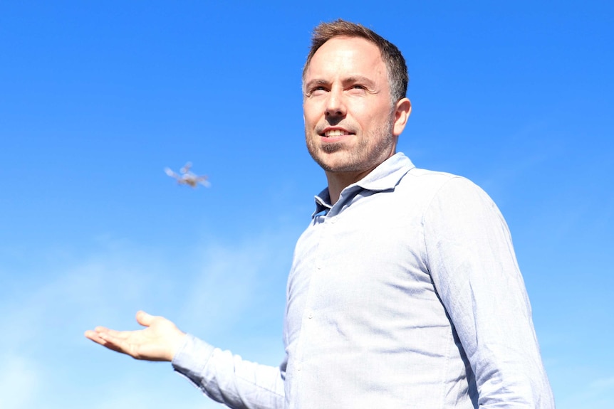 A man stands smiling into the distance as a drone flies in the sky behind him.