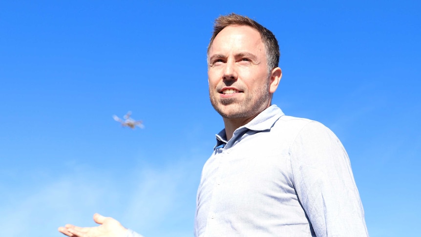 A man stands smiling into the distance as a drone flies in the sky behind him.