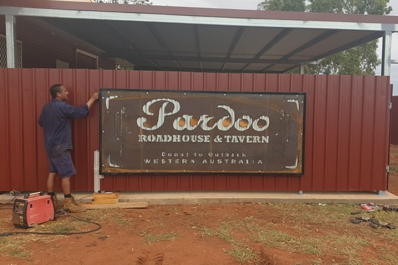 A man stands next to a large sign reading Pardoo roadhouse and tavern