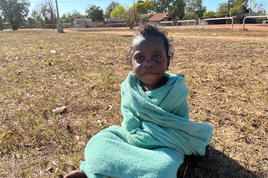 A young Indigenous child wrapped in a towel sitting cross-legged on grass.