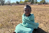 A young Indigenous child wrapped in a towel sitting cross-legged on grass.
