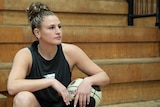 Basketballer Tiana Mangakahia sits in the stands of a basketball stadium, she looks into the distance, with a ball under her arm