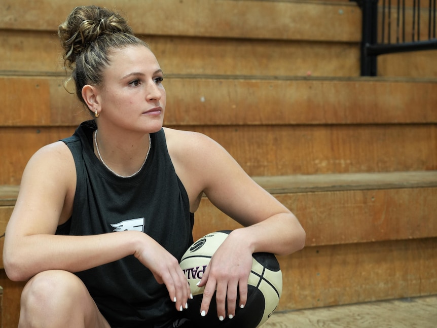 Basketballer Tiana Mangakahia sits in the stands of a basketball stadium, she looks into the distance, with a ball under her arm