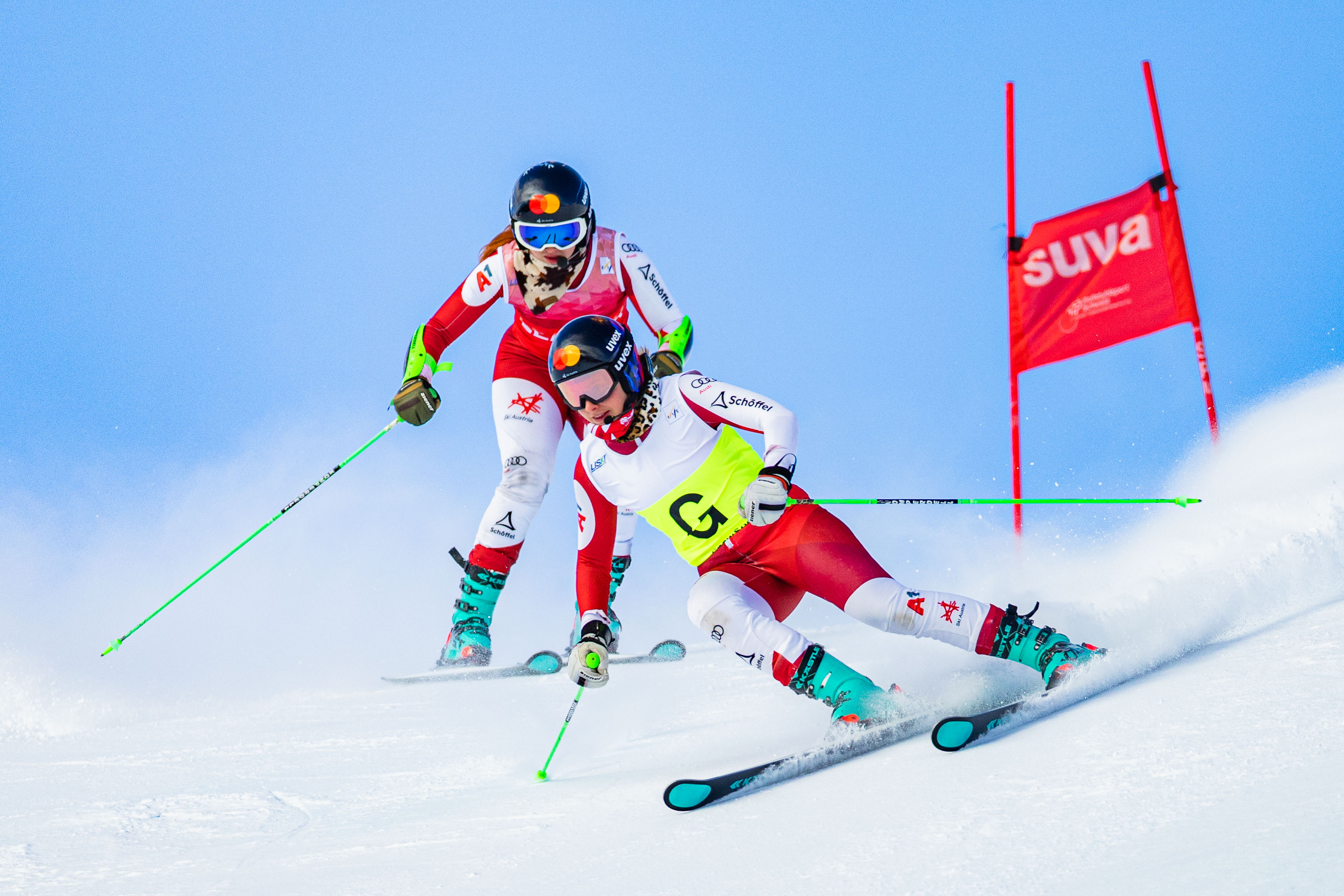 Two women ski around a flag and down a slope, while snow lifts from the ground to show how fast they’re going.
