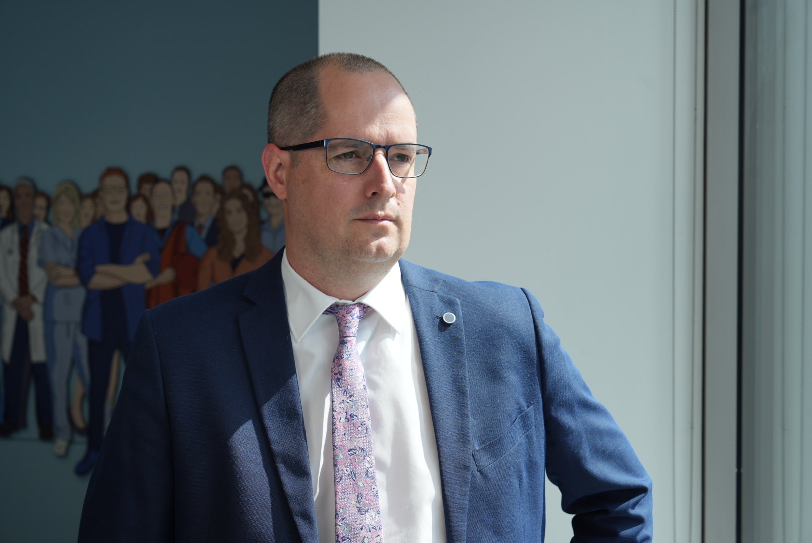 Shane Butcher in a navy suit, purple tie, glasses, in an office.
