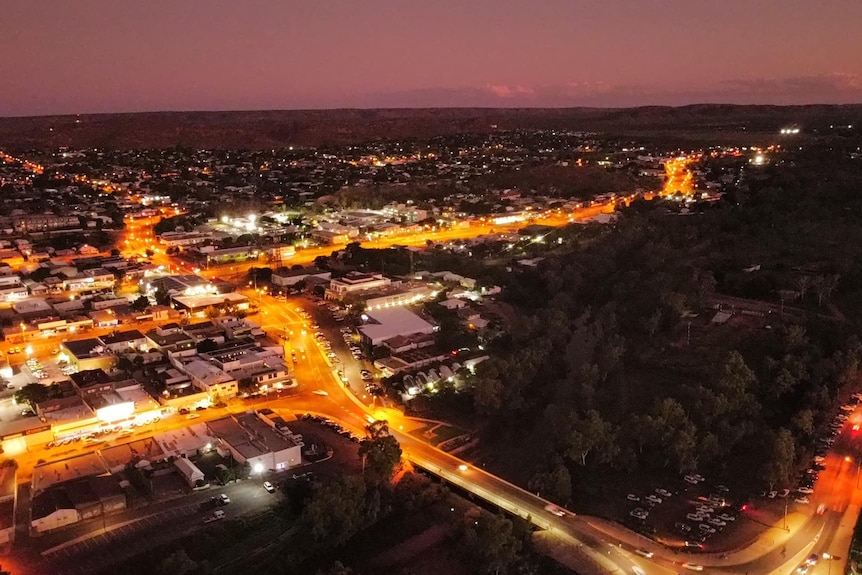An aerial view of an outback city at night with lit-up roads and houses and red dirt hills on the horizon