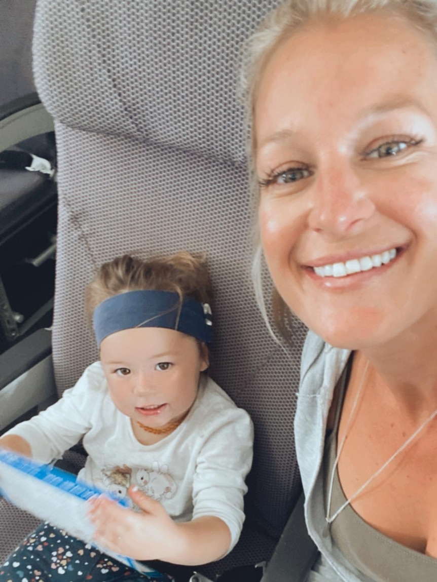 A blonde woman smiles with her daughter who holds an iPad in a chair. 
