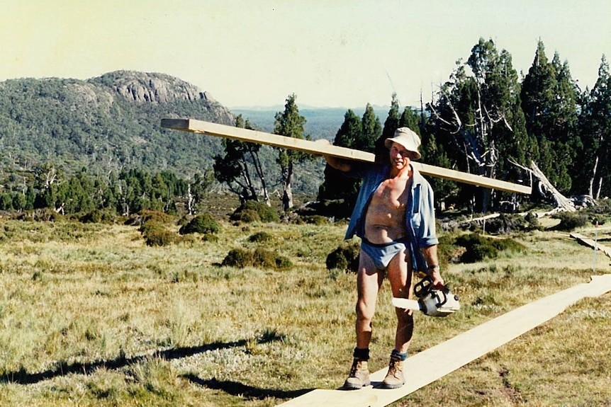 A man carries a plank of wood and a chainsaw along a wooden walkway in the mountains while wearing just undies and worker boots.