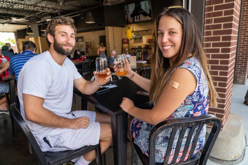 A young bearded man and a young woman with flowing dark hair with a band-aid on her shoulder hold their beer glasses together