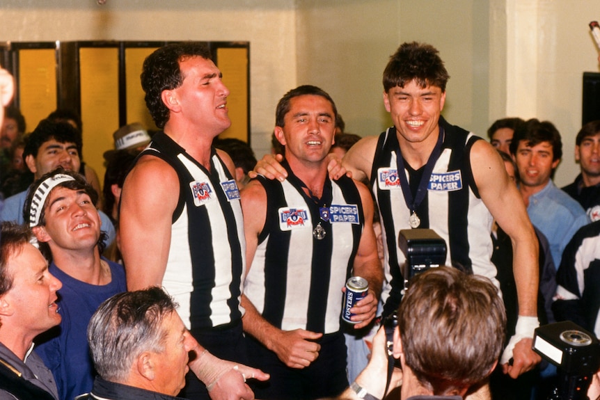 Darren Millane and Tony Shaw sing in the change rooms surrounded by lots of people.