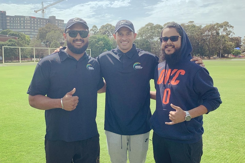 Three men pose for a picture on a cricket pitch in Sydney.