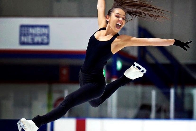 A woman in ice skates wearing all black jumps high above the ice.