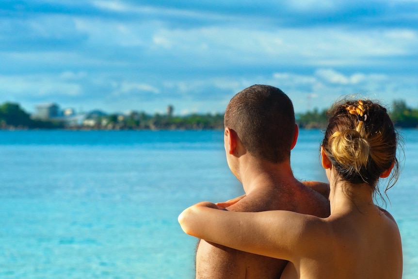 Looking out over turquoise water and trees beyond it, a couple is seen from behind, shoulders up, embracing.