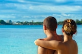 Looking out over turquoise water and trees beyond it, a couple is seen from behind, shoulders up, embracing.