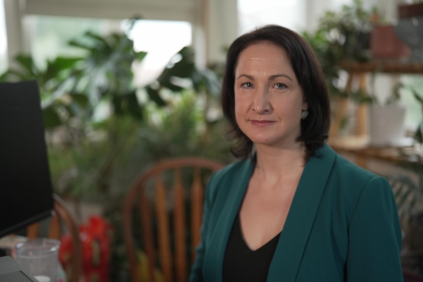 A white woman with brown hair wearing a green jacket. She is sitting in an office filled with plants