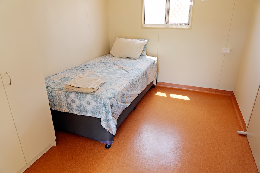 A small bedroom, featuring a single bed by a wall and a small window. 