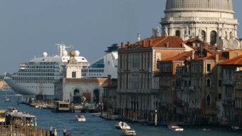 A cruise ship in the background is nearly as large as a large dome in Venice, while gondolas and vaporetto pass through a canal.