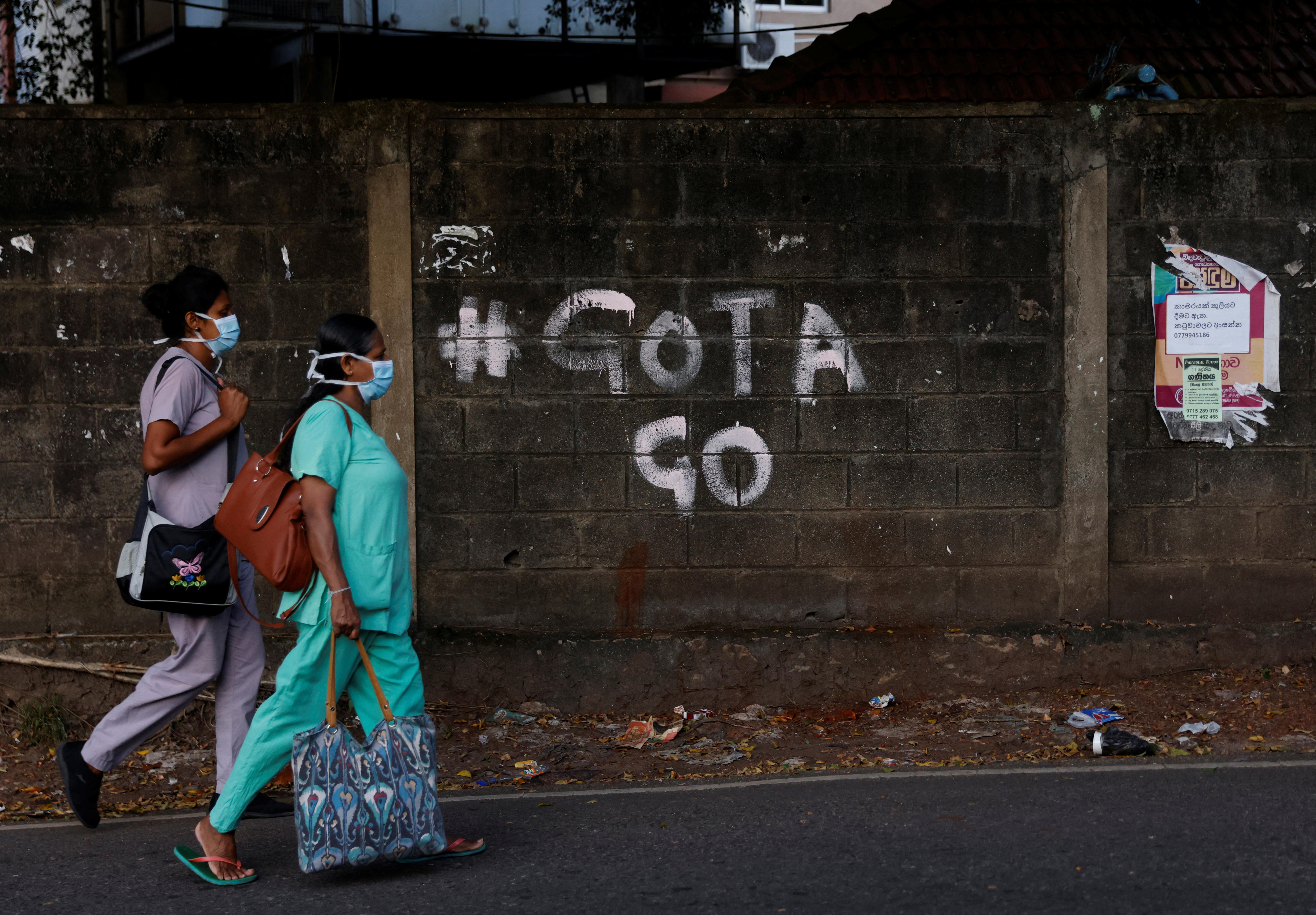 two workers in medical protective gear walk past grafitti on a wall reading "#GOTA GO"