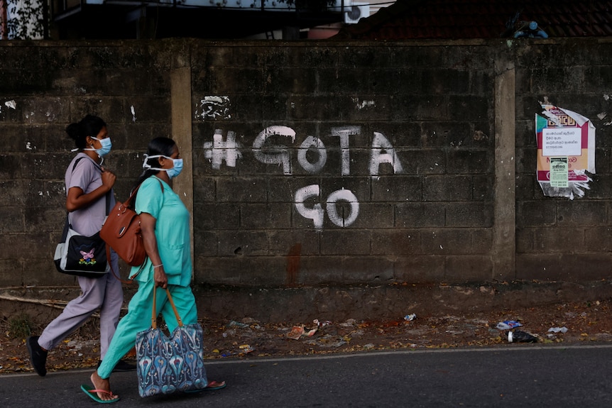two workers in medical protective gear walk past grafitti on a wall reading "#GOTA GO"