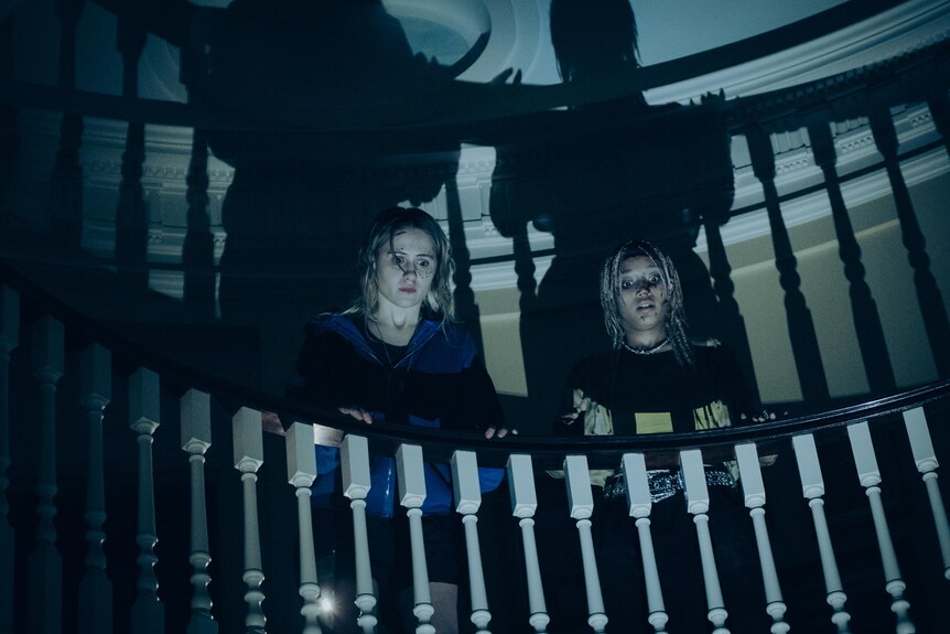 Two women stand in darkness on a staircase, with their shadows projected behind them on the wall.