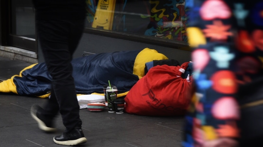 A homeless person is seen asleep in a sleeping bag on a footpath.