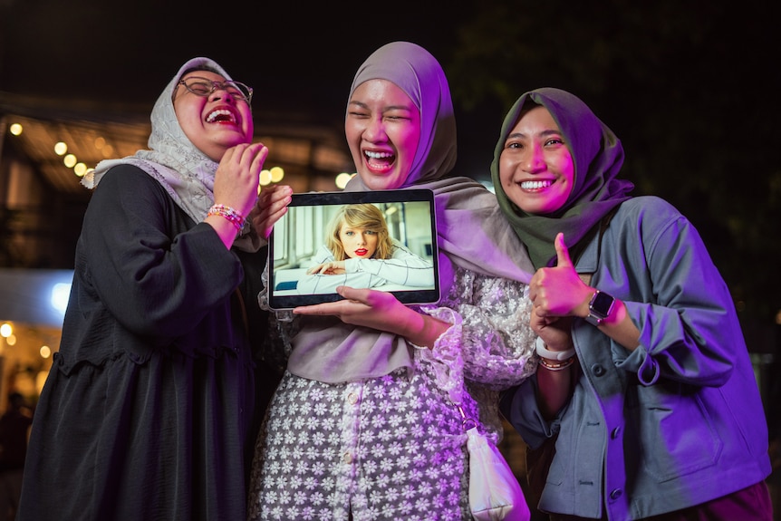 Three women wearing hijabs laugh as the woman in the middle holds an iPad with a portrait of Taylor Swift.