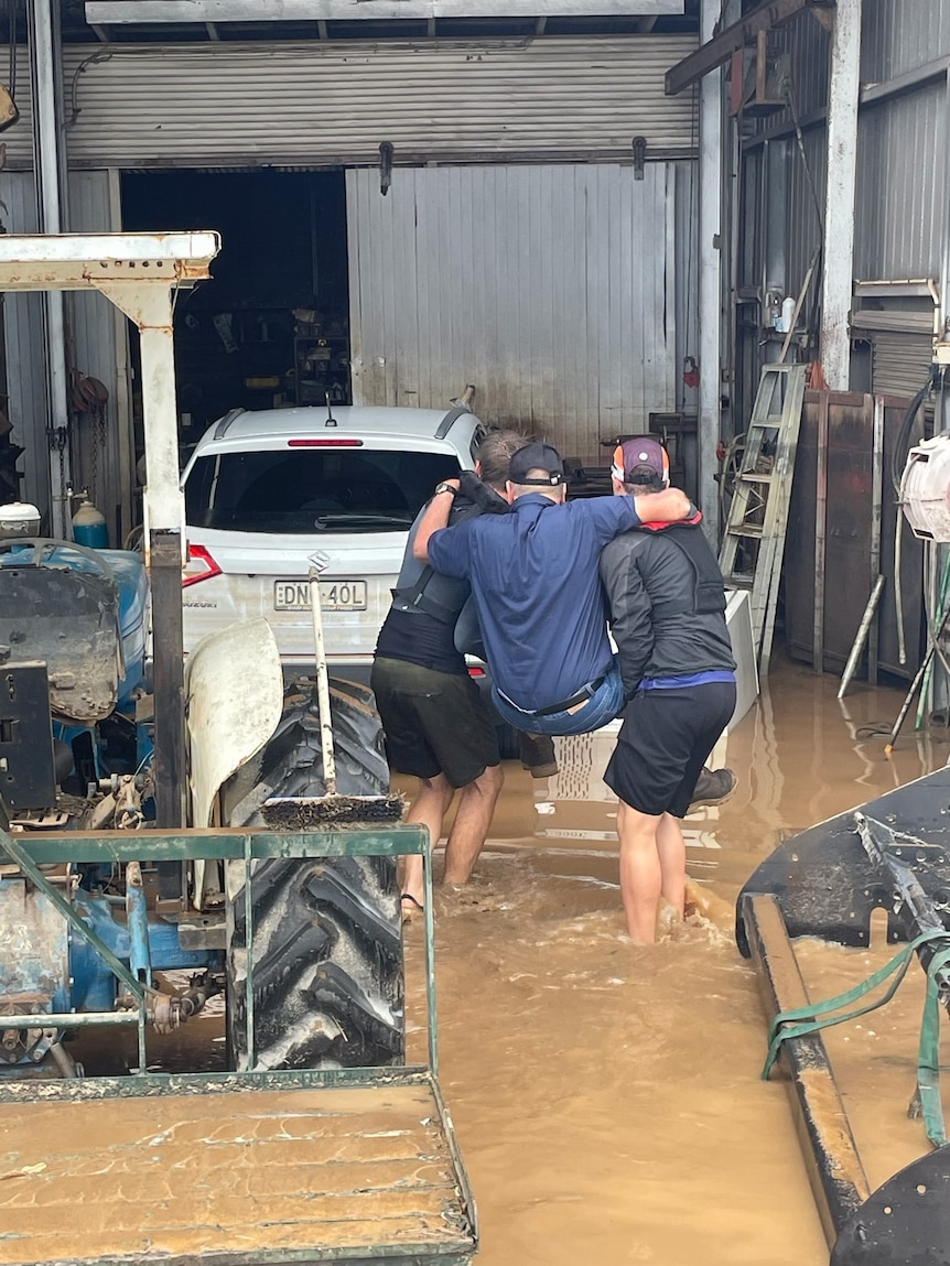 Two men in black shorts carrying another man through a flooded workshop.