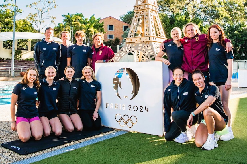 A group shot of swimmers and coaches around a Paris 2024 sign at a swimming pool.