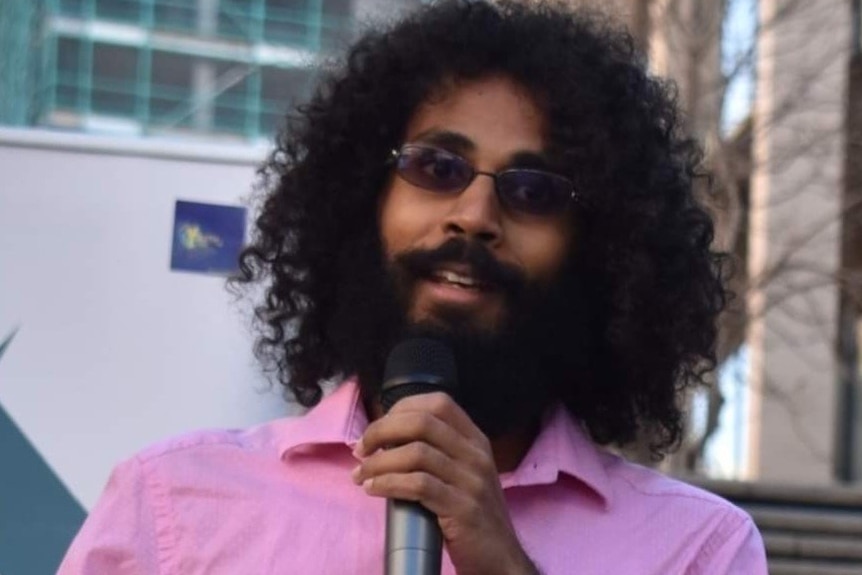 A man wearing a pink shirt speaks into a microphone at a rally.