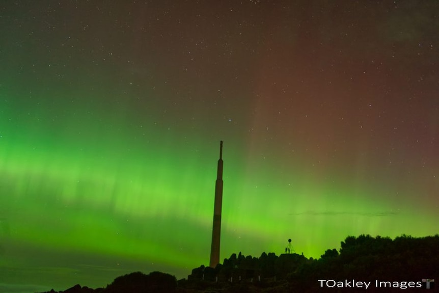 Lights of aurora australis over a mountain summit with large antenna pole.