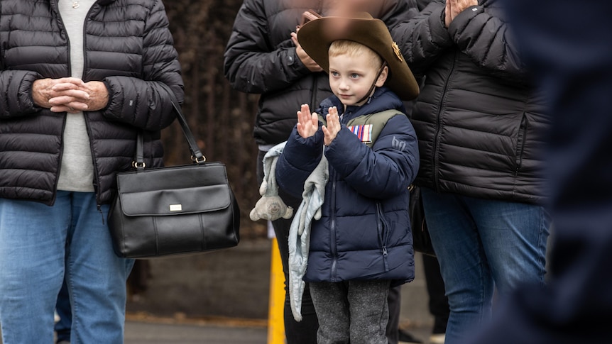 A small boy wearing a warm coat and medals stands among adults, watching the march.