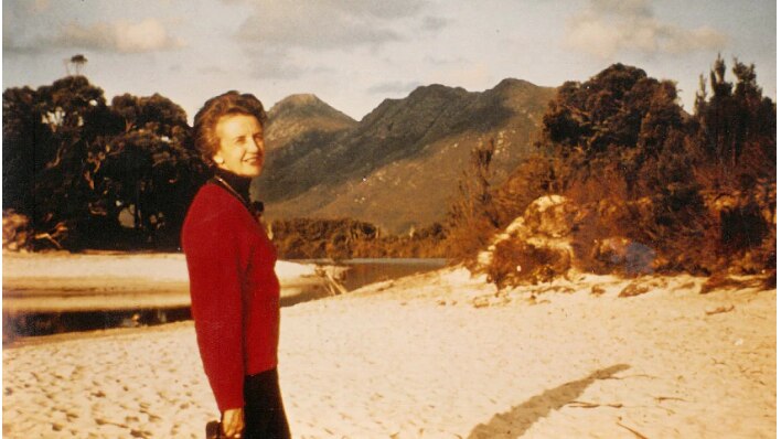 Old photo of a woman wearing a red sweater standing on a beach with mountains in the background.