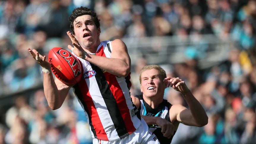 An AFL player launches himself in the air with his hands grabbing for the ball ahead of a defender.