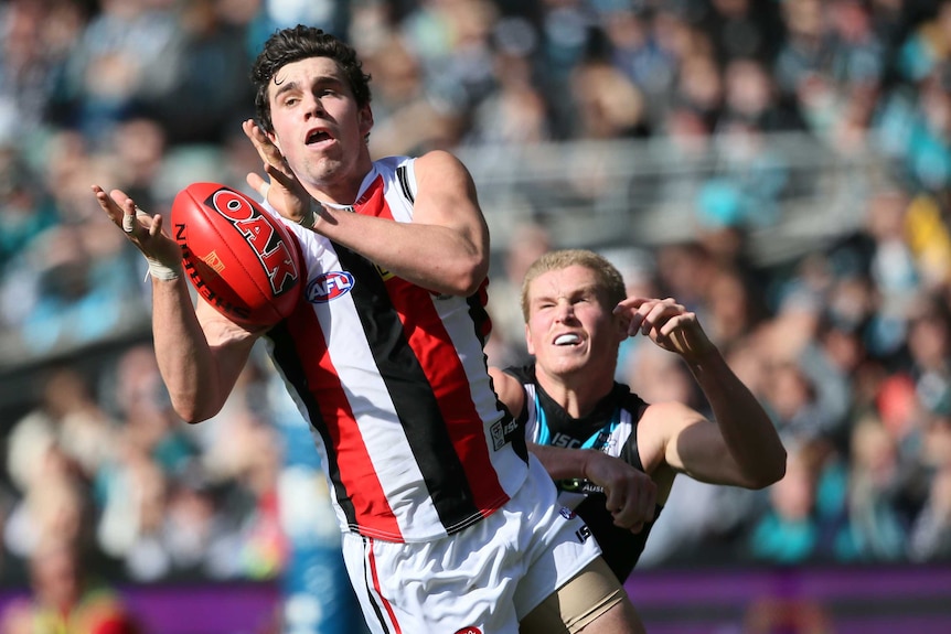 An AFL player launches himself in the air with his hands grabbing for the ball ahead of a defender.