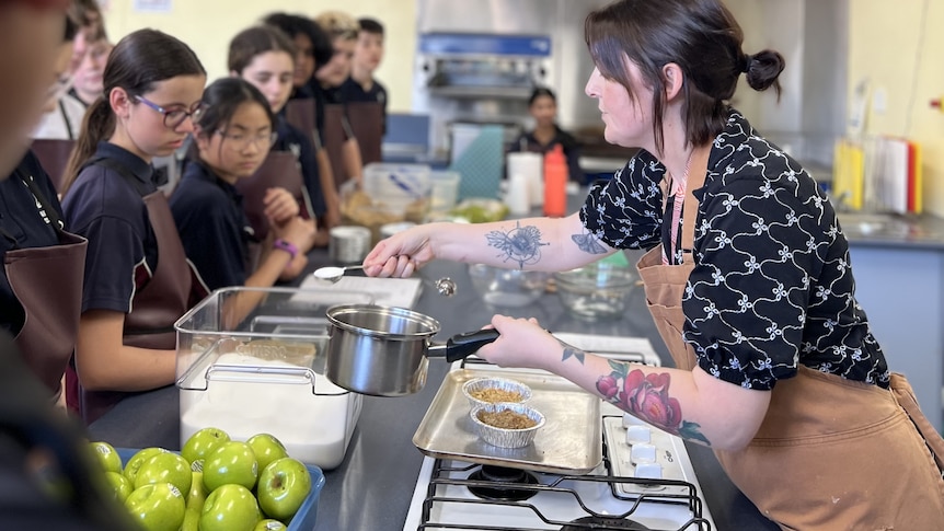 High school students observing female teacher demonstrating ingredient measurement in cooking class.