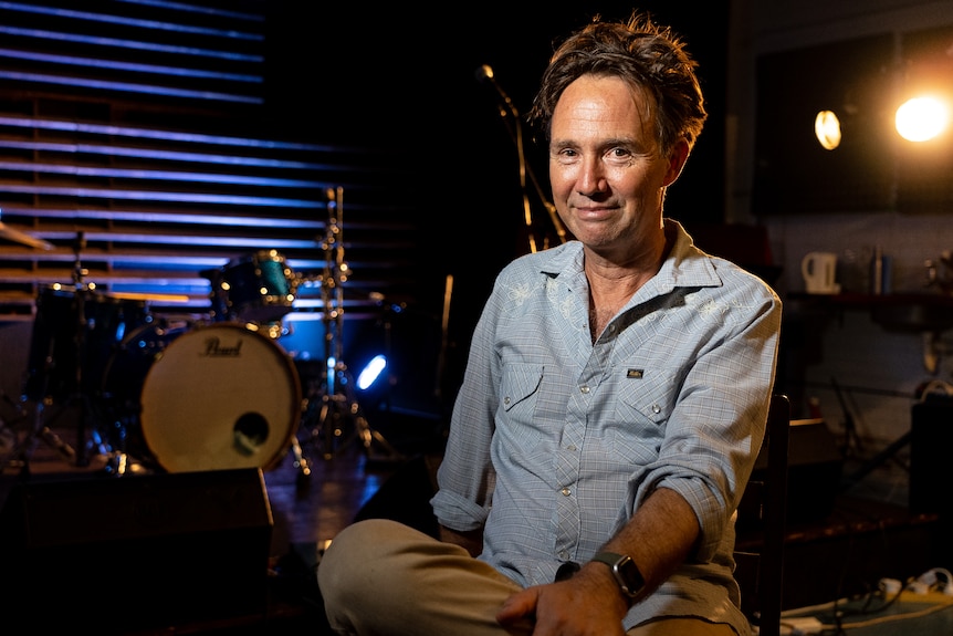 A middle-aged man sits in a darkened music studio, a drum-kit behind him