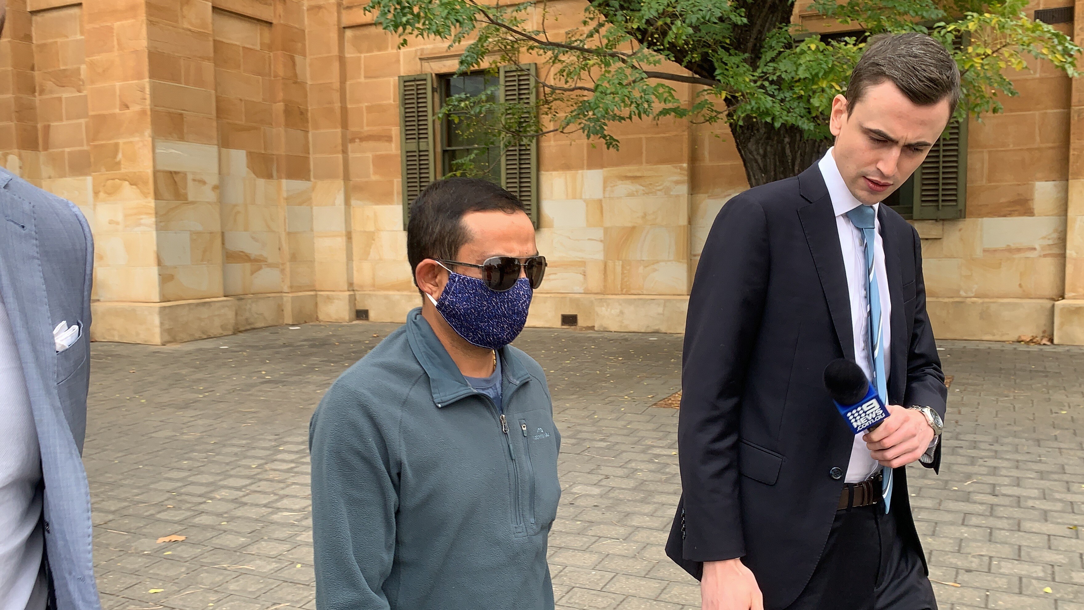 A man with short dark hair wearing sunglasses and a blue spotty face mask 