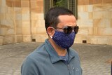 A man with short dark hair wearing sunglasses and a blue spotty face mask 