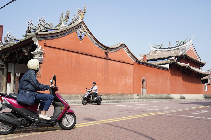 two scooters drive past peach building.