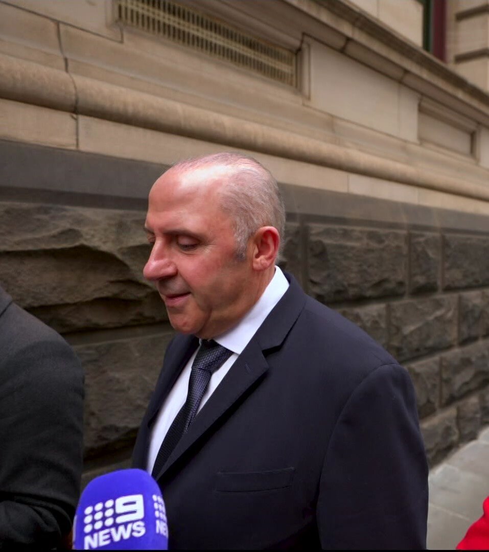 A balding man with wisphs of white and grey hair in a dark suit walks beside a stone building, looking down.