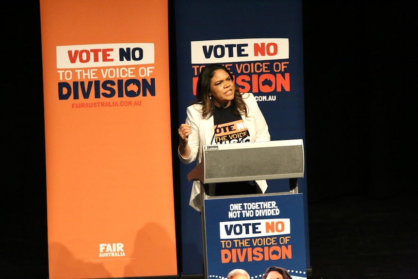 A woman wearing a white jacket speaks at a lectern. 