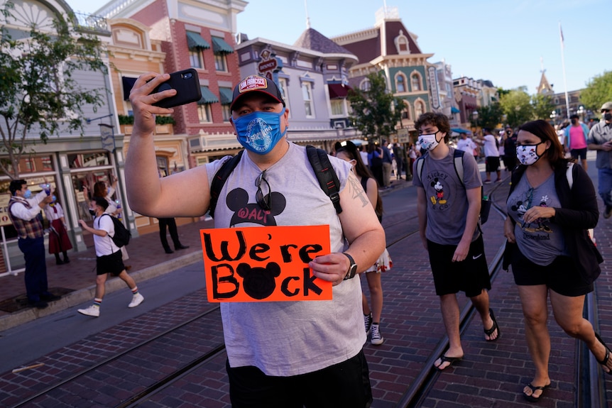 A man in a mask walks with a crowd at Disneyland holding a sign saying "We're back".