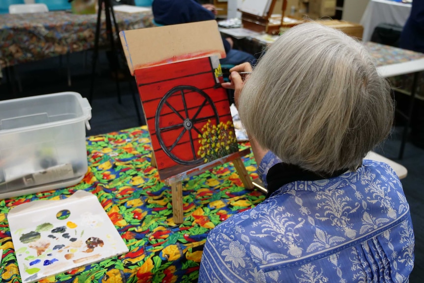 A grey haired woman paints a wagon wheel on a red background.