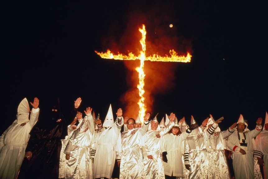 Klu Klux Klan members wearing hooded robes gesture as they burn a cross.