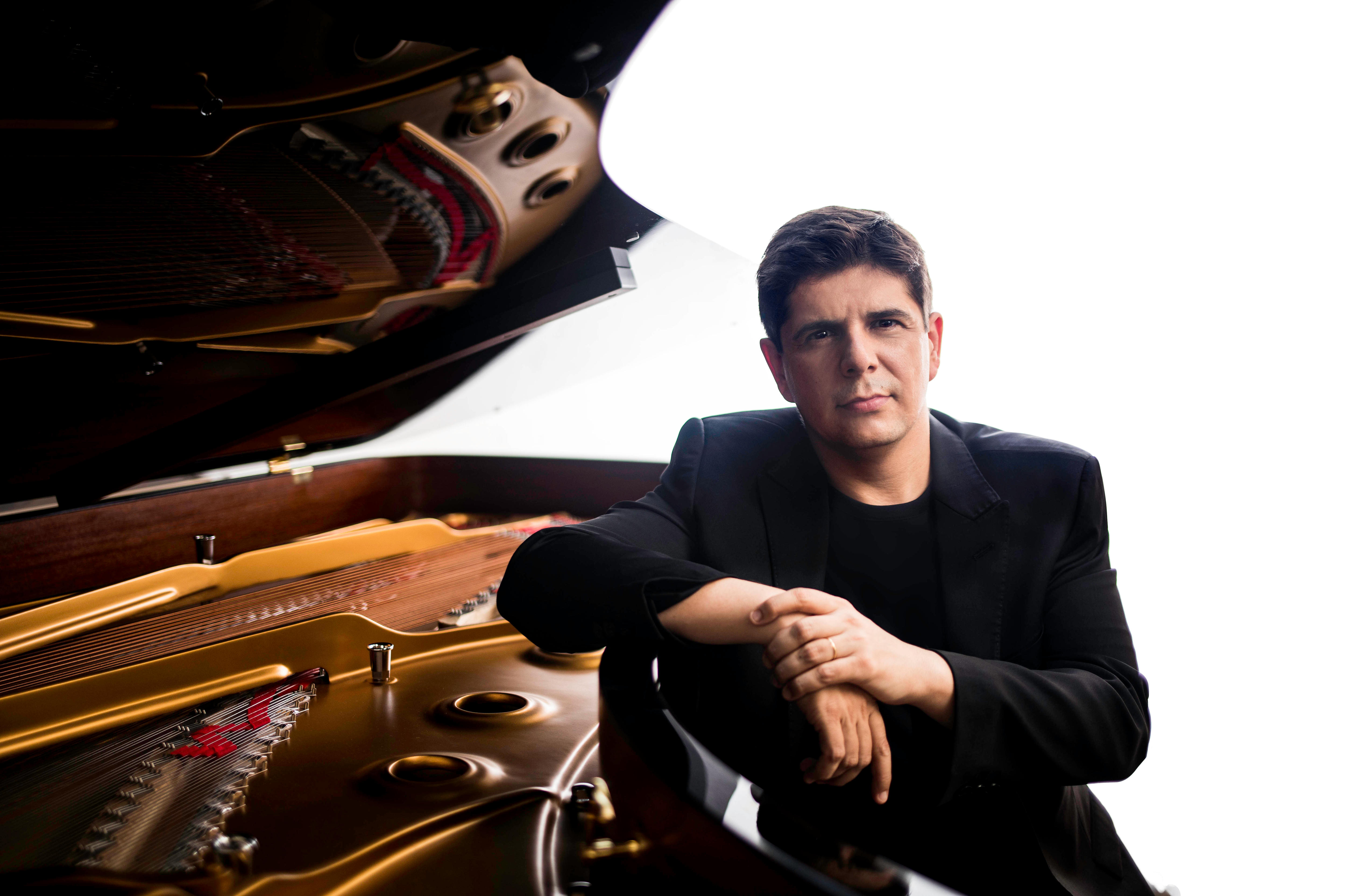 Javier Perianes sits at an open grand piano, with his arm resting up on the body. He is wearing a black jacket and shirt.