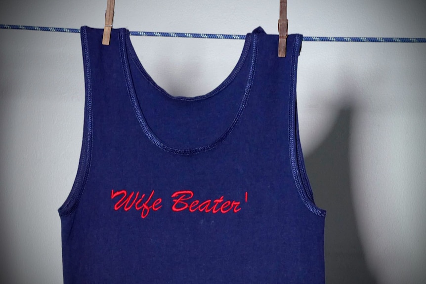 A blue singlet hanging by pegs on a clothes line, embroidered with the words 'wife beater'.
