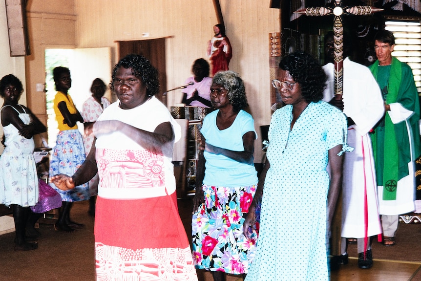 Aboriginal women dance inside a church. They are wearing colourful clothing and have their arms painted in white 
