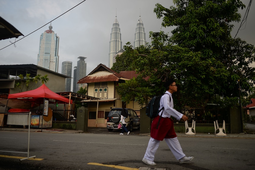 A boy in traditional white attire walks past a wooden home. The Petronas Towers loom in the background.