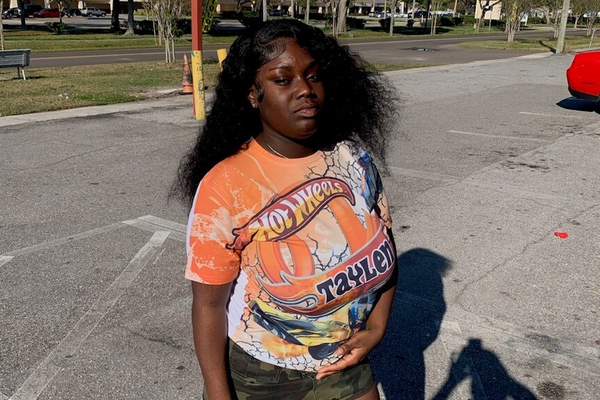 A young black woman with long hair stands in a concrete parking lot wearing a Hot Wheels t-shirt.
