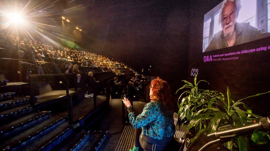 Man with grey beard on a large Imax screen with a large crowd in theatre and woman with red hair and green jacket at front